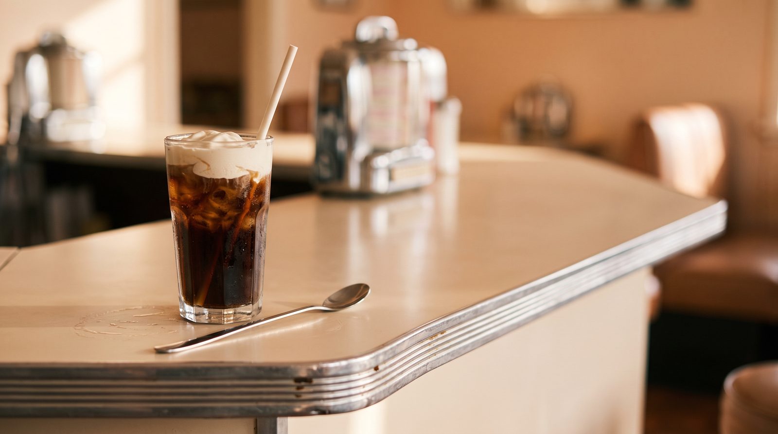 Tall glass of cola over ice with a cream cap, a long spoon and a paper straw on a 1950s-style milk-bar counter